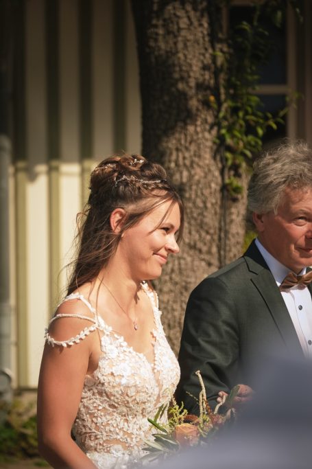 Braut in klarem Spitzenkleid, begleitet von ihrem Vater. Die Tränen halten sich noch zurück