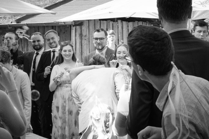 Eine Gruppe von Personen feiert den ersten Kuss des Brautpaars. Alle sind umgeben von festlicher Dekoration im Freien.